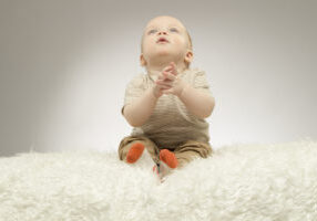 Adorable little baby sitting on the white blanket in praying pose, studio shot, isolated on grey background,
